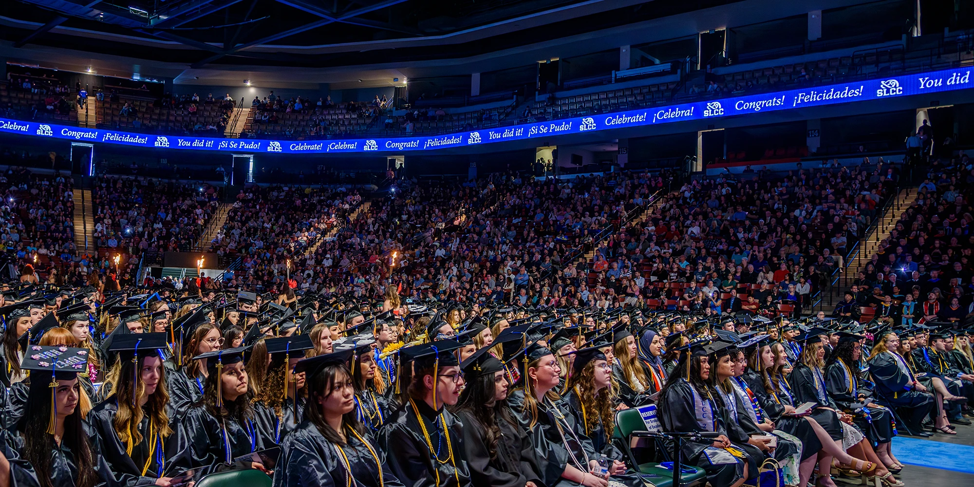 Large indoor graduation ceremony with rows of graduates in caps and gowns seated in the foreground, facing a stage. Audience members are seated in the stands. An electronic banner overhead displays congratulatory messages including 'Congrats!', 'Felicitades!', 'You did it!', and 'Celebrate!'