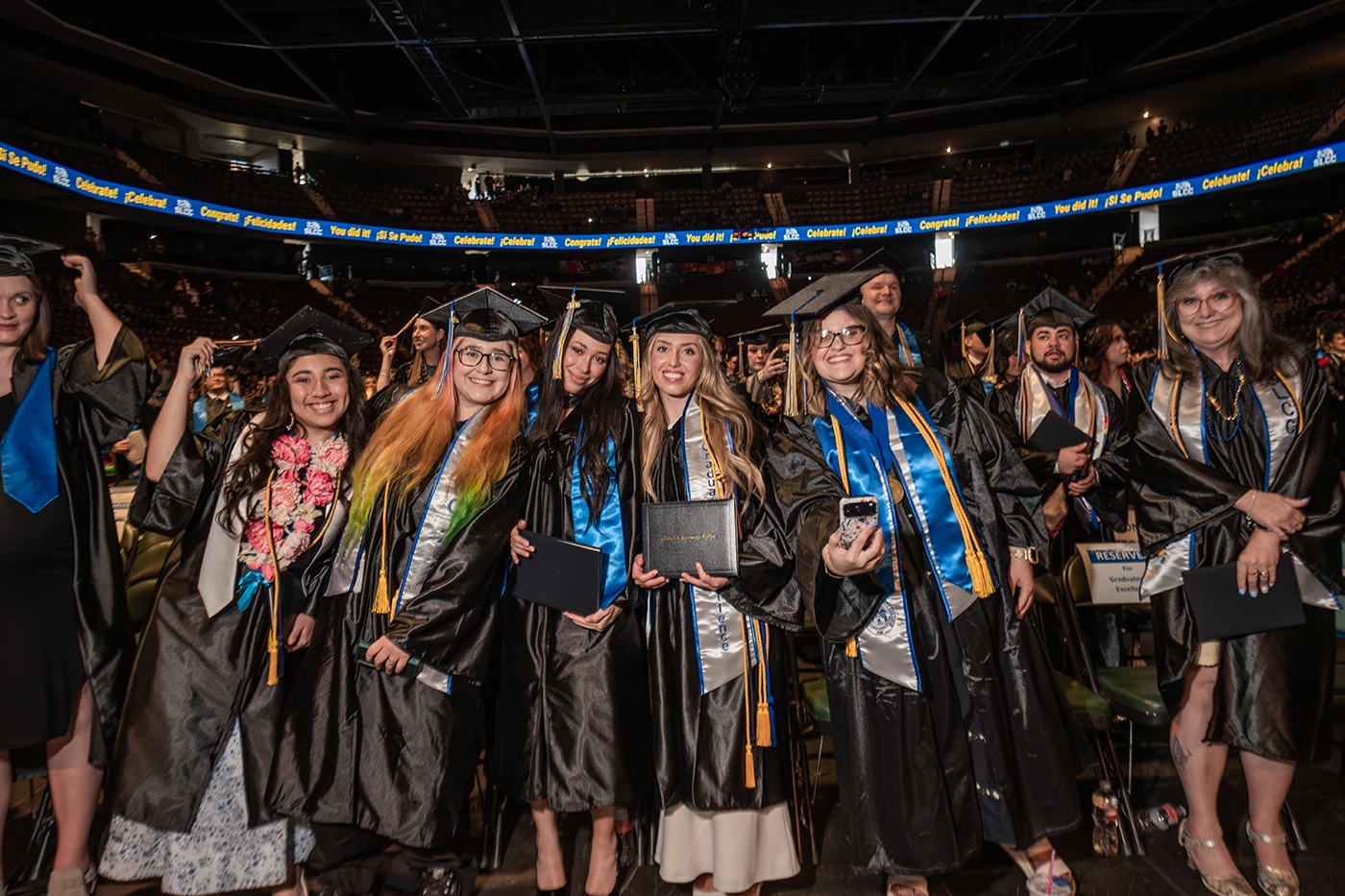 A group of graduates in black caps and gowns smile and pose together at a commencement ceremony inside a large arena. Several hold diploma covers and wear blue and gold honor stoles.