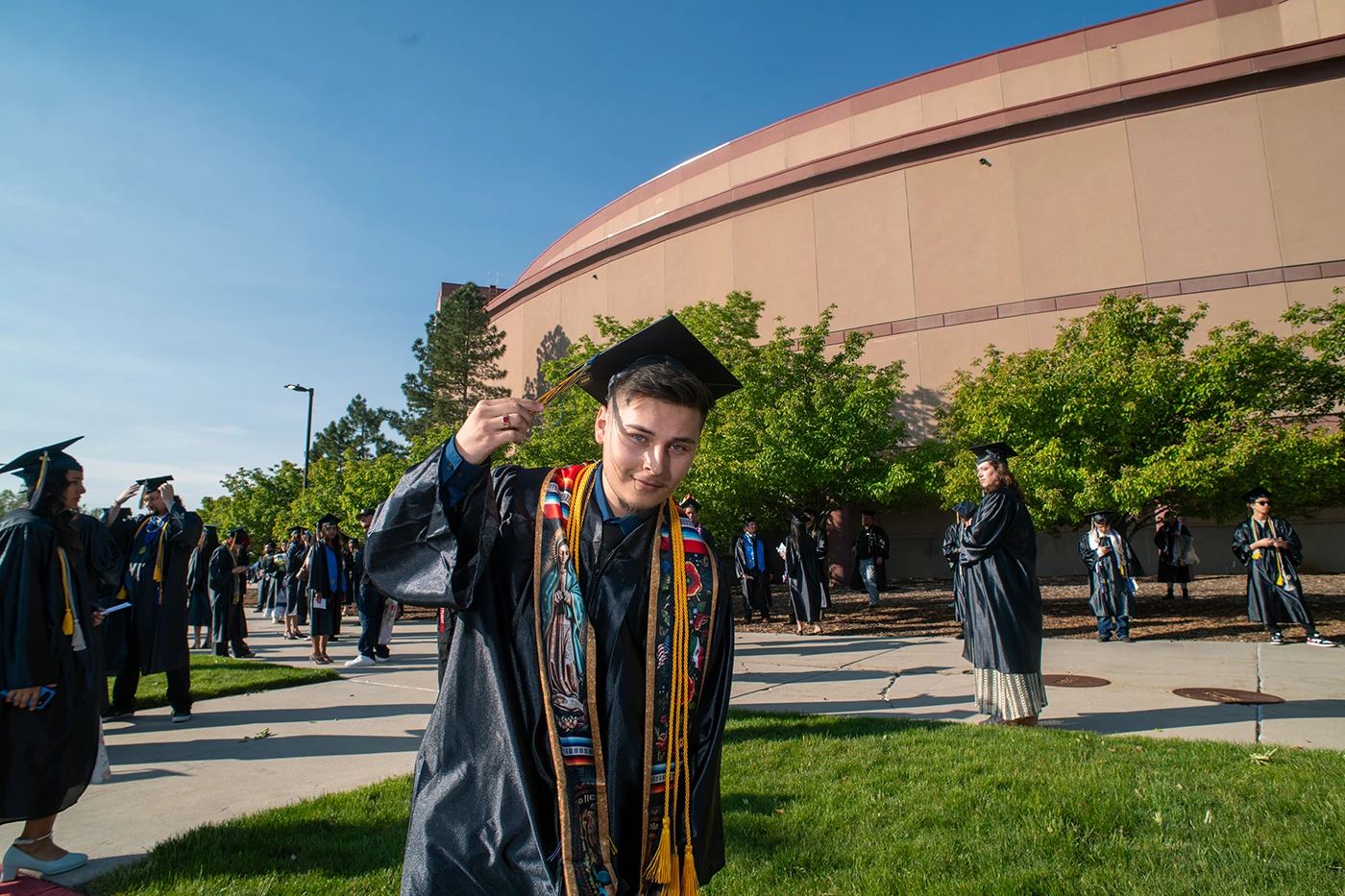 Graduate in cap and gown holds up tassel before commencement ceremony, colorful embroidered stole visible, other graduates and arena building in background.