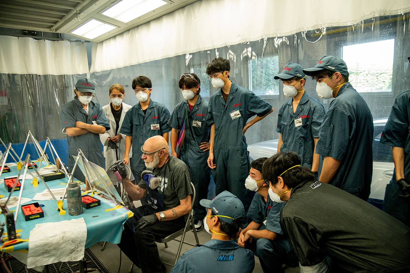Instructor demonstrating airbrush painting techniques to a group of students wearing protective coveralls, safety glasses, and respirators in a training workshop.
