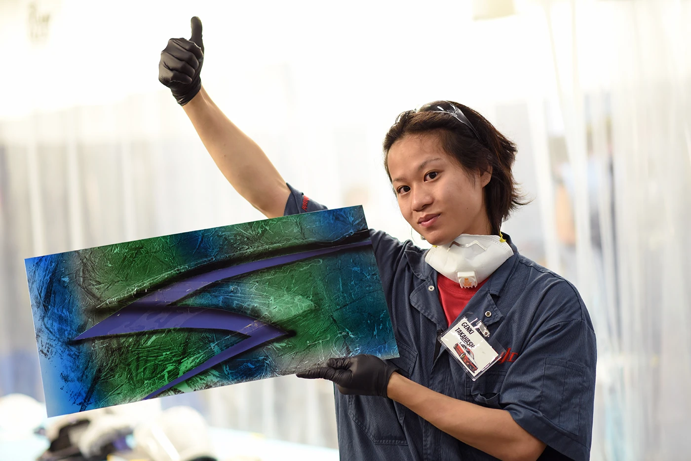 Person in a workshop wearing protective gloves and a respirator around the neck, holding a painted panel with a blue and green abstract design and giving a thumbs-up.