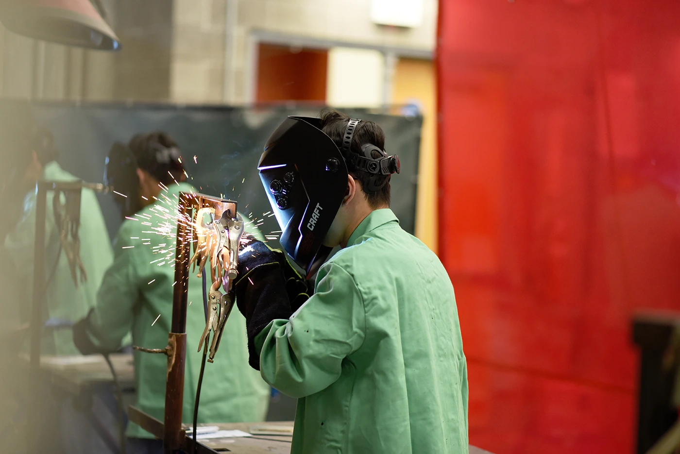 Person wearing a welding helmet and protective clothing performing a weld on a metal frame, with sparks visible at the point of contact.