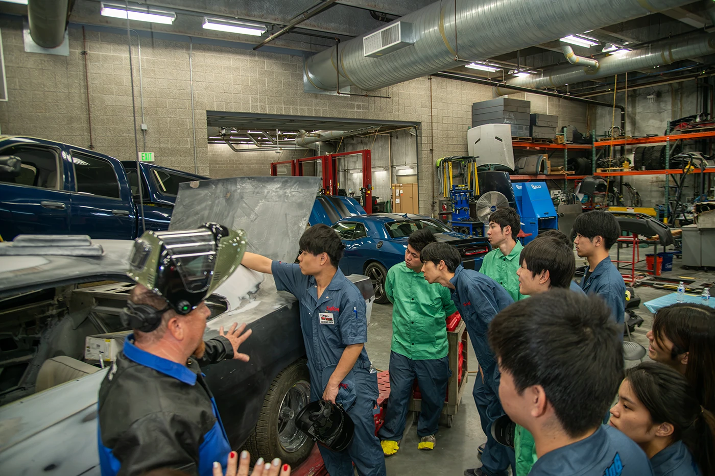 Instructor and students gathered around a vehicle in an automotive workshop, discussing body repair techniques beside a partially restored truck.