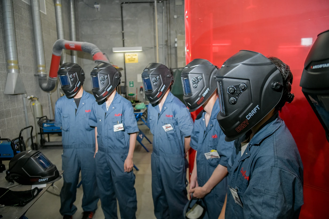 Students wearing welding helmets and protective coveralls stand in a welding lab during a safety briefing or demonstration, with welding equipment and ventilation ducts visible in the background.