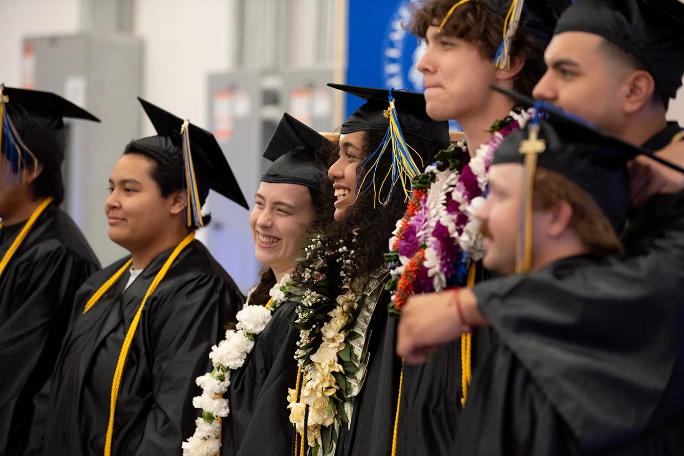 Several students lined up wearing black graduation regalia, mortarboards, and brightly colored floral leis.