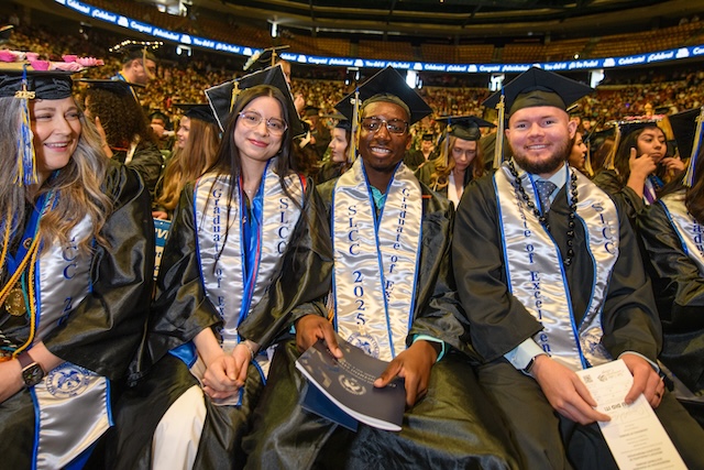 Group of graduates seated in a large auditorium or stadium, wearing caps, gowns, and sashes with text such as 'SLCC 2025.' The background shows a large crowd attending the ceremony.