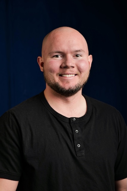 A smiling bald man with a trimmed beard, wearing a black henley shirt, posed against a dark blue background in a professional portrait.