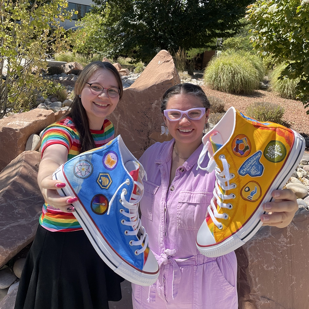 Two students hold shoes towards the camera with red rocks and green foliage behind them. One student wears a rainbow-striped shirt with a black skirt. The other wears a pastel purple jumpsuit with matching glasses. The shoes are bright blue and bright yellow-orange with white laces and soles, and they’re painted with small icons.