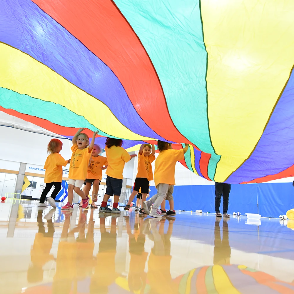 Young children wearing matching orange shirts play underneath a large, colorful parachute in a gymnasium at The Eccles Early Childhood Development Lab School. The bright parachute reflects on the polished floor as the children smile and interact.
