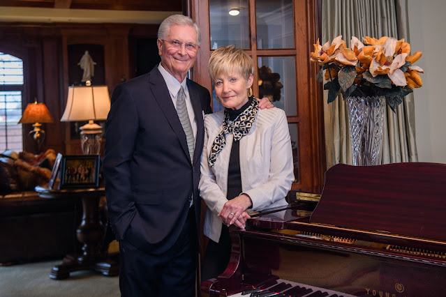 An older man in a suit and an older woman in a light jacket and patterned scarf stand closely together, smiling beside a polished grand piano in a warmly lit living room with wood-paneled walls and floral décor.