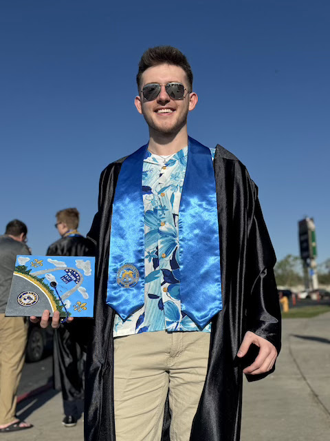 Person wearing a graduation gown and blue stole with an emblem, holding a decorated graduation cap. Other graduates are visible in the background. The photo is taken outdoors under a clear blue sky. 