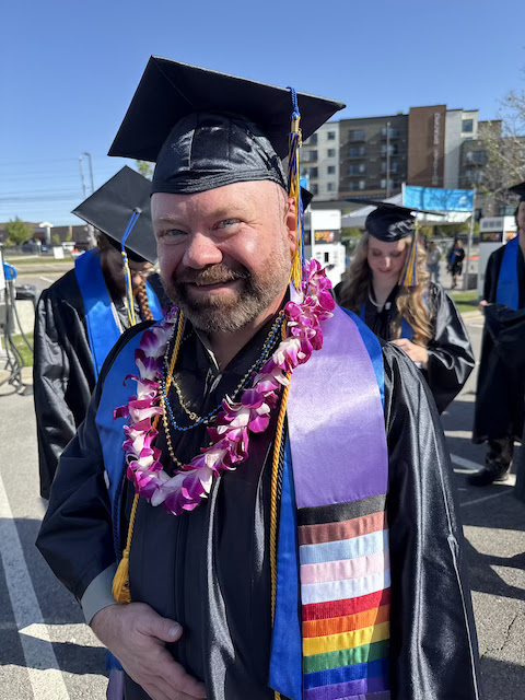 Person in graduation cap and gown wearing a purple lei and a stole with the LGBTQ+ pride flag. Other graduates are visible in the background.