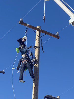 Two workers climbing a utility pole, wearing safety harnesses and helmets, working on electrical lines. Clear blue sky in the background.