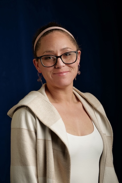 A woman with dark hair pulled back, wearing glasses, a light pink headband, and dangly earrings, smiles gently. She is dressed in a cream and beige plaid hoodie over a white top, posed against a dark blue background in a professional portrait.