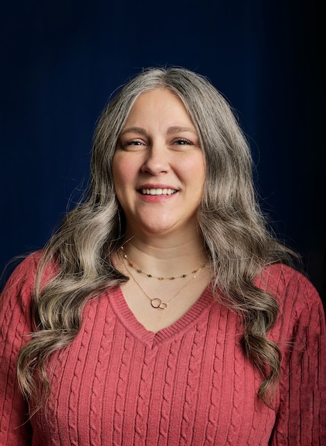 Person with long, wavy hair wearing a red cable-knit sweater and two necklaces, standing against a dark blue background.