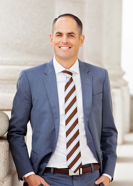 A man in a blue suit with a brown and white striped tie stands smiling against a stone column in a formal, classical architectural setting.