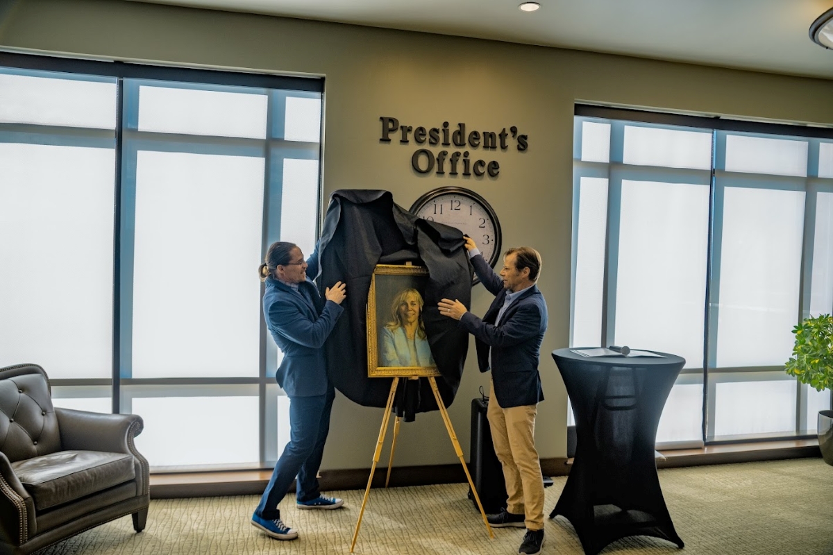 Middle-aged man, wearing a blue suit with blue converse tennis shoes, with red hair, beard, glasses and ponytail, helps artist Rob Adamson unveil a painting of former SLCC President Huftalin
