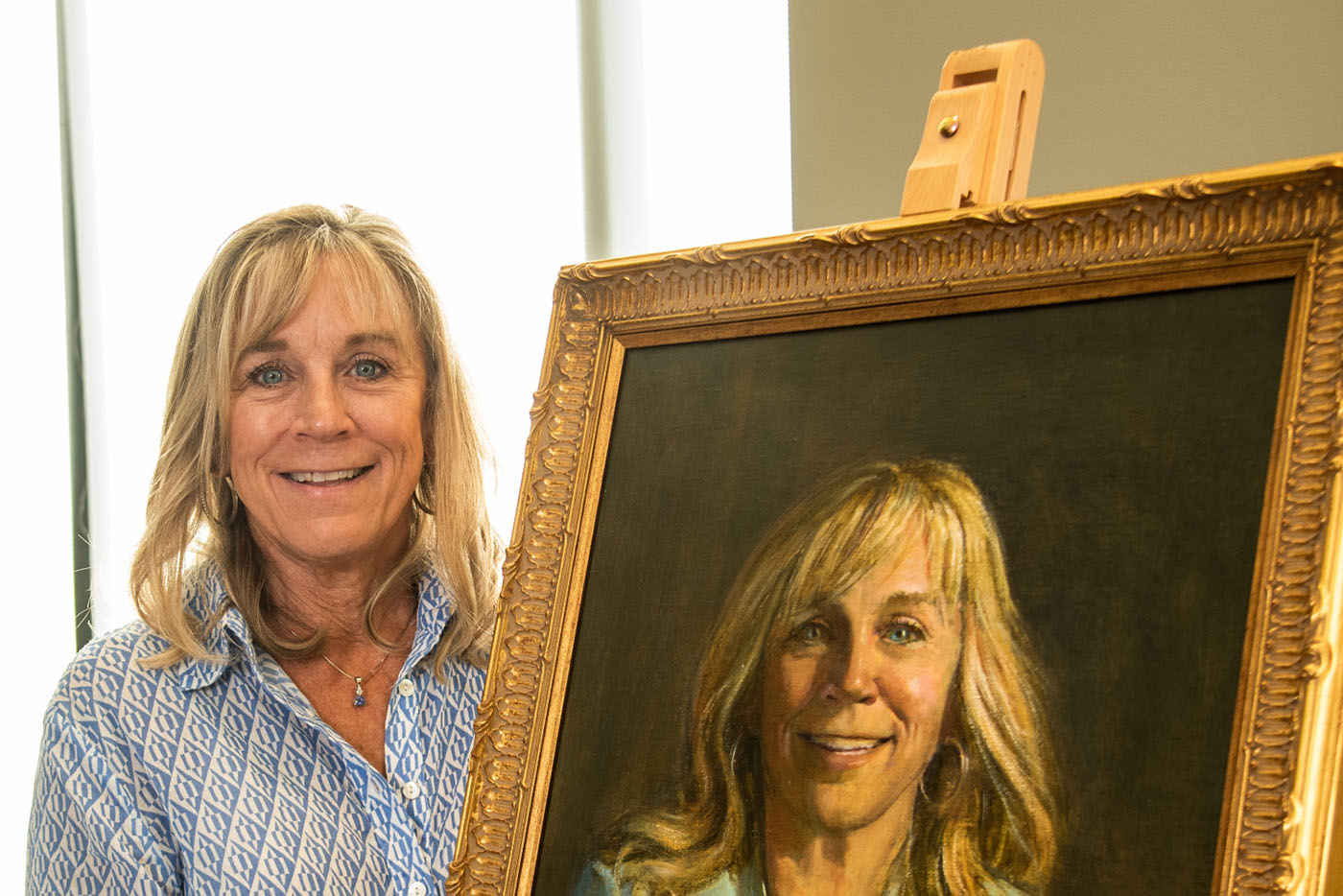 A blond middle aged woman stands next to a painting of herself.