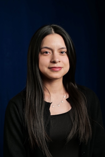A woman with long, straight dark hair smiles softly. She is wearing a black top with a black cardigan and a delicate necklace with small star-shaped charms. The background is a deep blue, and the photo is a professional portrait.