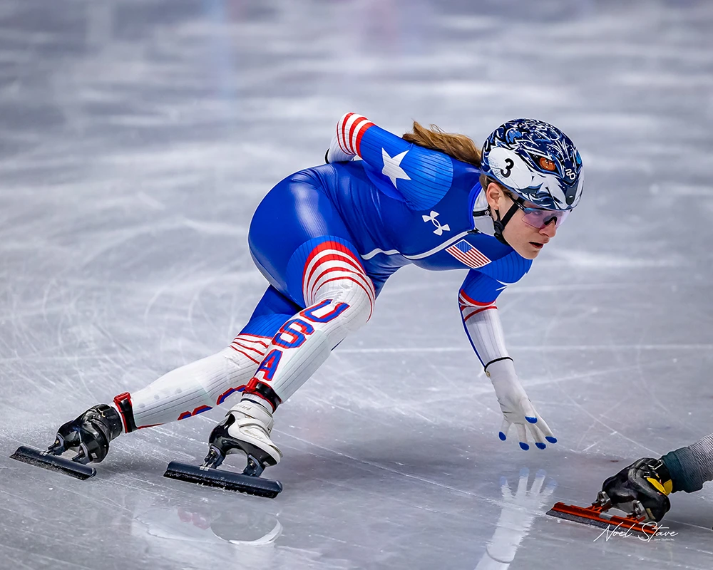 A speed skater cornering hard, leaning over and dragging her inside hand on the ice. She's wearing a blue and white team USA skinsuit with a stylized wolf head painted on her helmet.