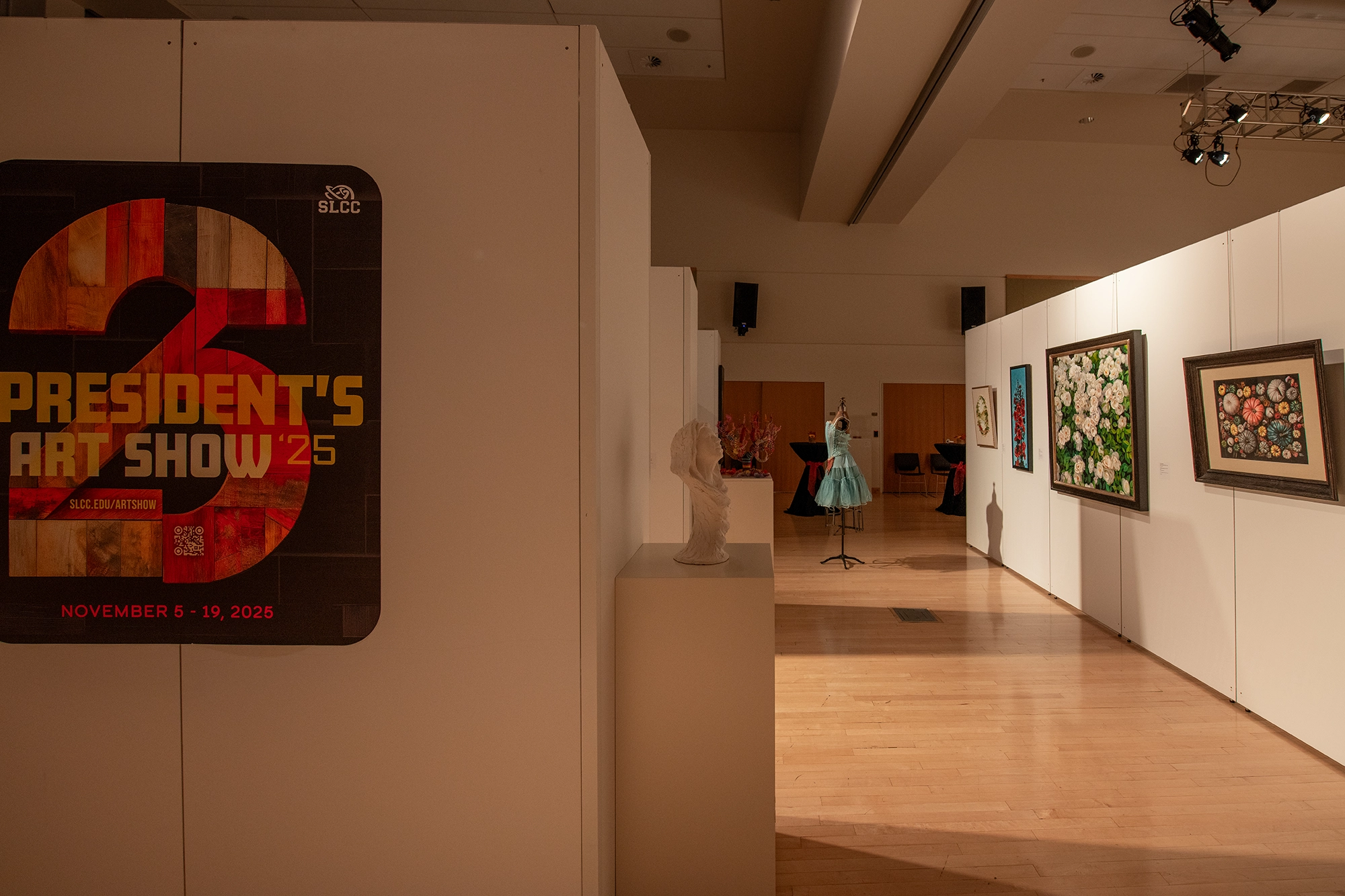 Art exhibition space with white partition walls displaying framed floral paintings, a white sculpture bust on a pedestal, and a mannequin in a blue gown. A large poster on the left promotes the SLCC President's Art Show 2025, with event dates and a QR code. The gallery has wooden floors and overhead lighting, creating a professional and welcoming atmosphere.