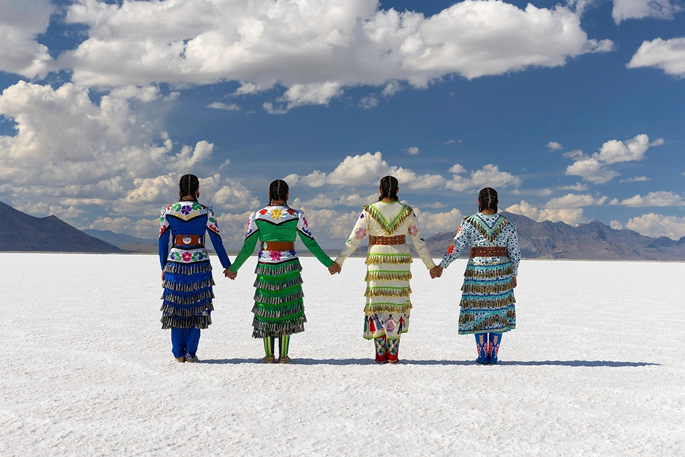 Photograph. Four American Indian women wearing brightly colored and tasseled dresses and standing in a line facing away from the camera. The women are holding hands, creating a striking symmetry in the center of the photograph. Bright white salt flats stretch to the horizon, just below the centerline of the photograph, and the distant background is brown and purple mountains beneath a blue sky with well-defined cumulus clouds.