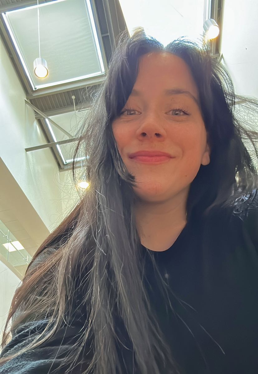 The artist, smiling in a selfie with long brunette hair and wearing a long-sleeved black sweatshirt.