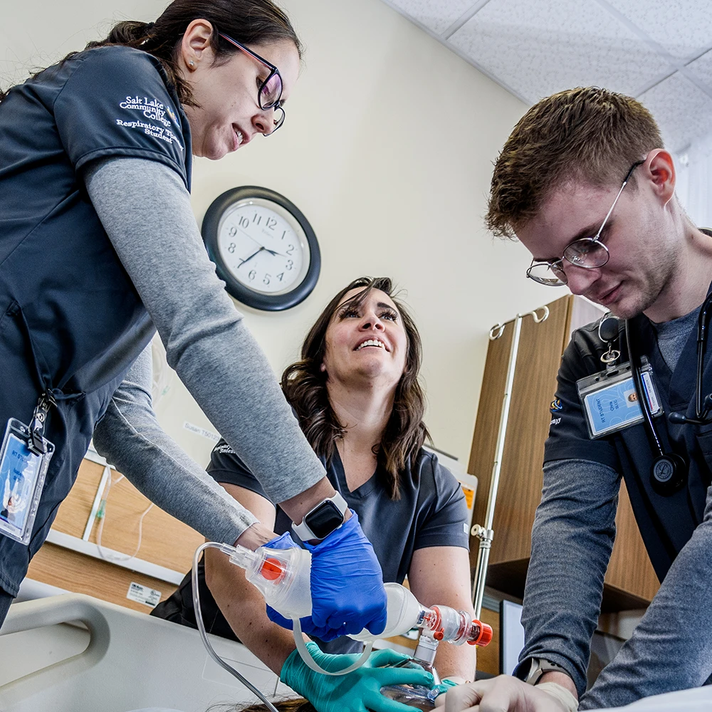 Three people in medical scrubs work together in a clinical simulation at SLCC’s Health Science Respiratory Therapist Program. One operates a manual resuscitator on a patient mannequin while the others assist. They are in a hospital lab setting with medical equipment and a clock on the wall.