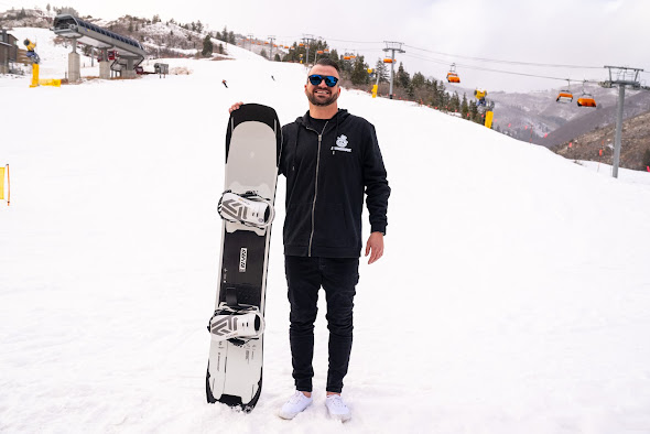 Person standing on a snowy slope holding a snowboard vertically. Wearing a black jacket with white text and logos, black pants, and white shoes. Ski lifts with orange seats and snow-covered hills are visible in the background.