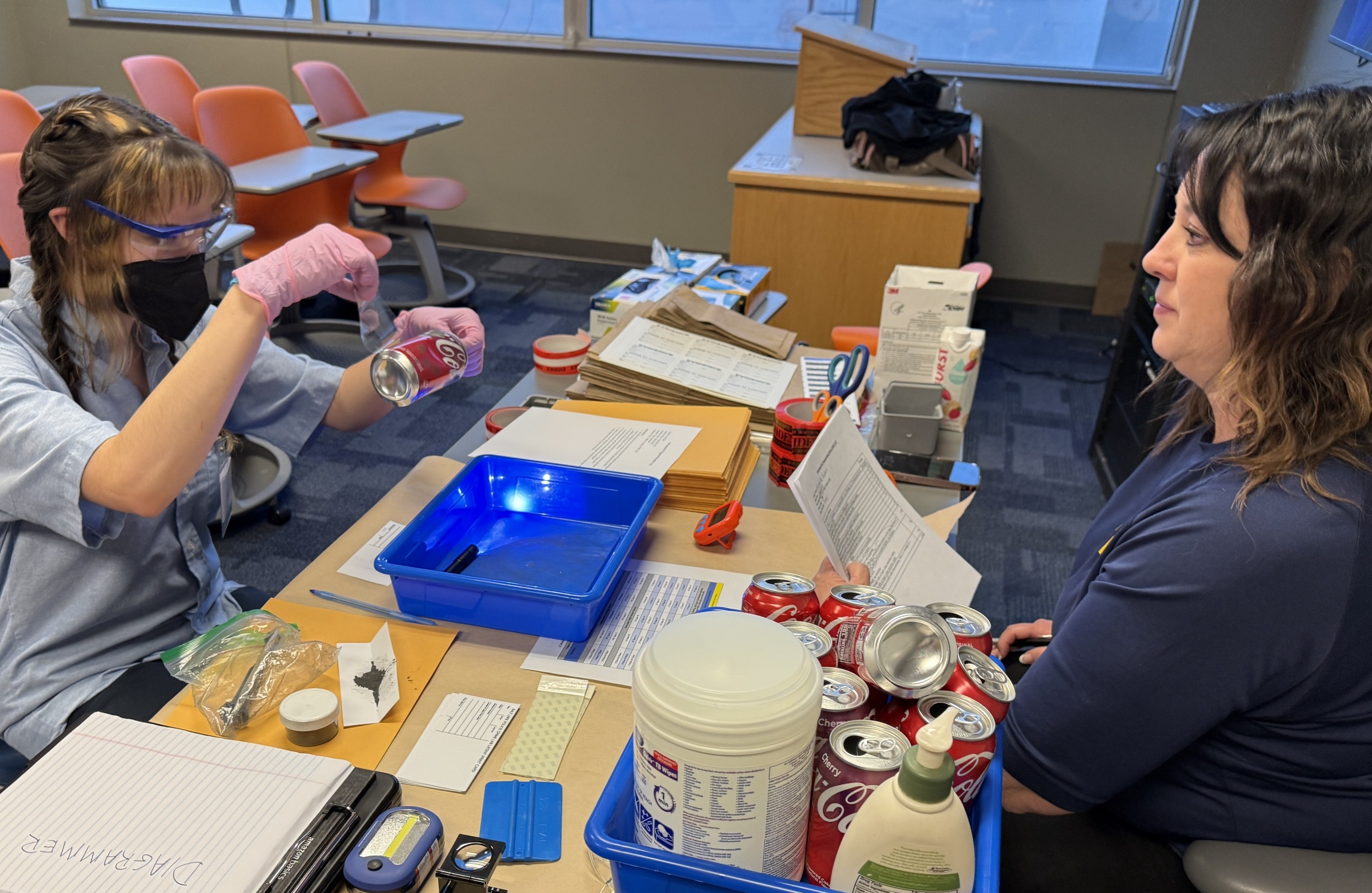 SLCC student wearing safety glasses and gloves participates in a SkillsUSA Crime Scene Investigation competition while an advisor observes at a classroom table filled with evidence materials.