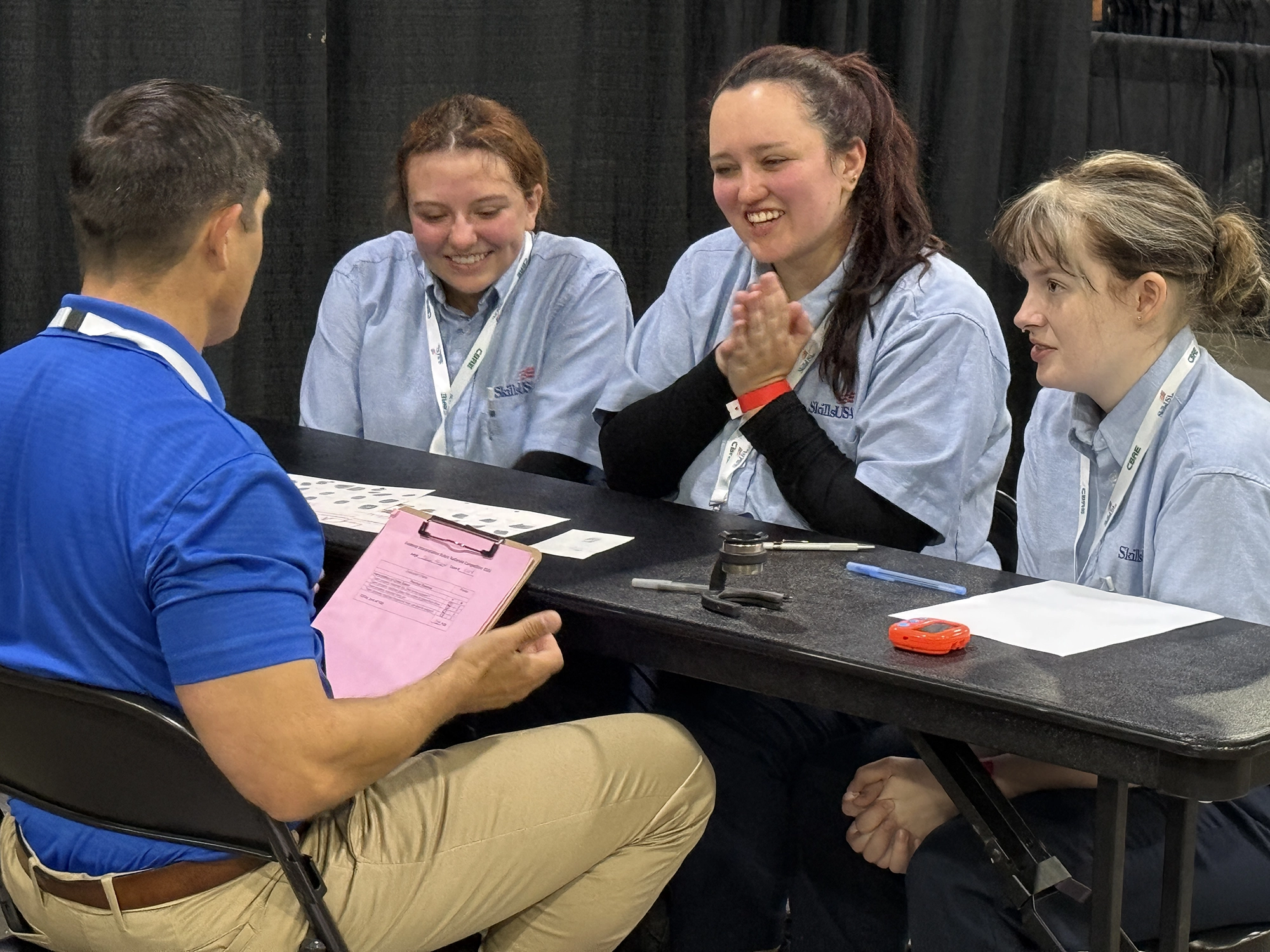 SLCC Forensic Science Students, left to right, Kennedi Leon, Haylee Ramirez and Kylee Hermeling sit at a table and answer questions from a judge, a man with short black hair with back to camera, at the National Skills USA competition.