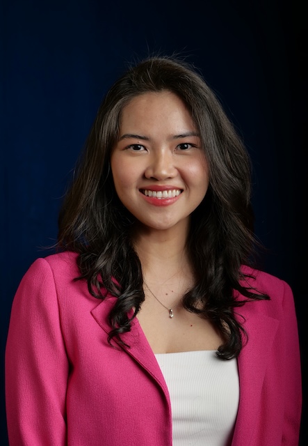 Person with long, dark hair wearing a pink blazer over a white top, standing against a dark background.