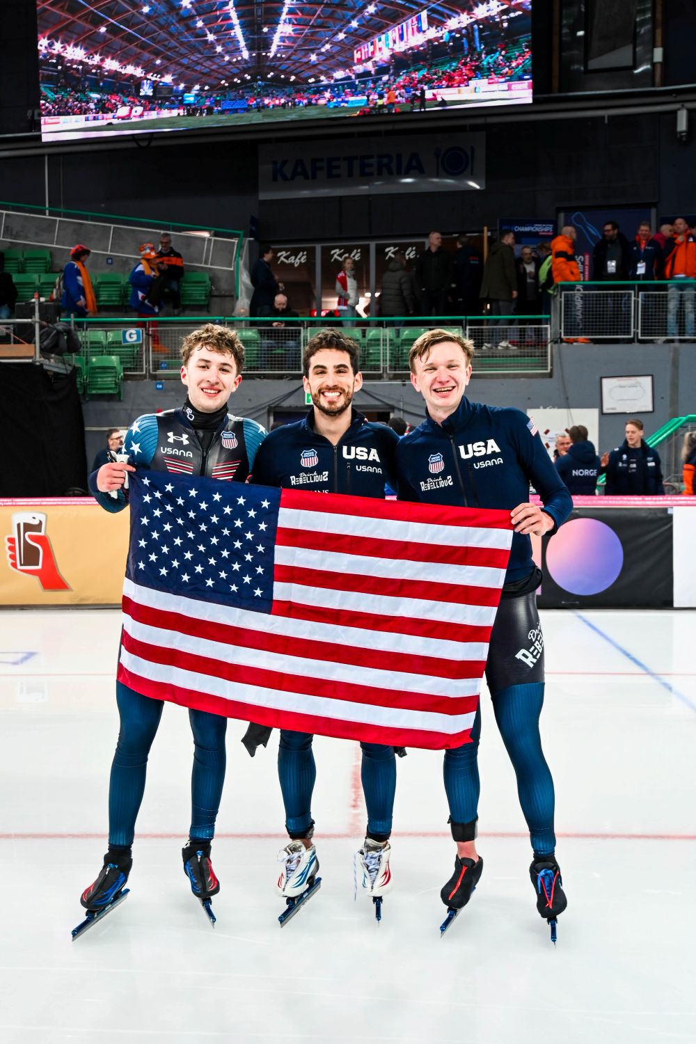 Three U.S. speedskating athletes stand on the ice in an arena, smiling and holding an American flag after a world championship win.